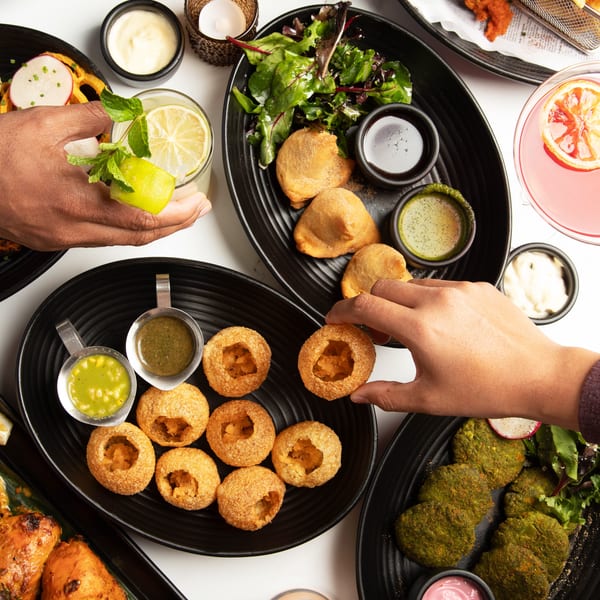 Overhead view of various Indian dishes, including pani puri, samosas, fried chicken, and drinks, with hands reaching for food.