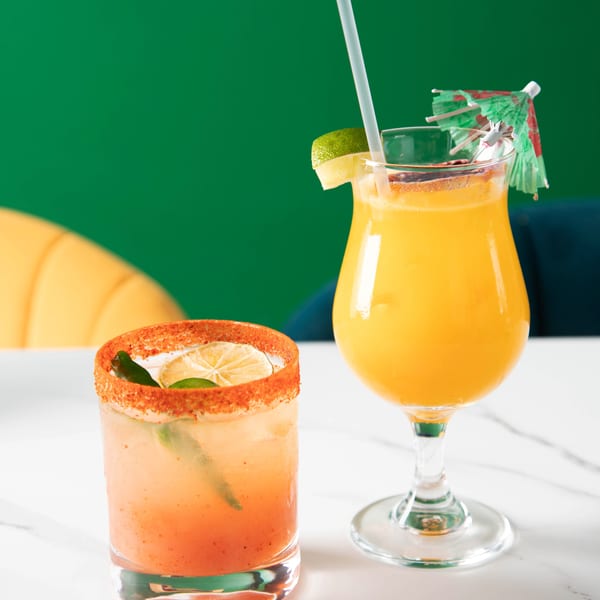Two vibrant cocktails, one orange with a chili-lime rim, the other yellow with a lime and umbrella.