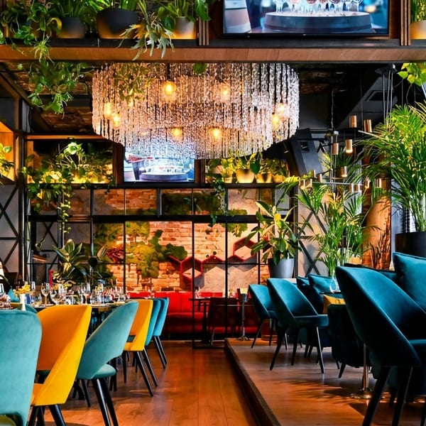 A modern restaurant interior with teal and yellow chairs, lush green plants, and a large crystal chandelier.
