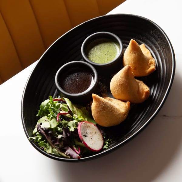 Three golden samosas, two dipping sauces, and a side salad with radish on a black plate.