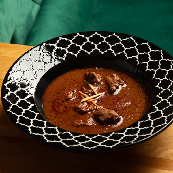 A black bowl with white geometric patterns holds a rich brown curry with meat pieces and ginger slivers.