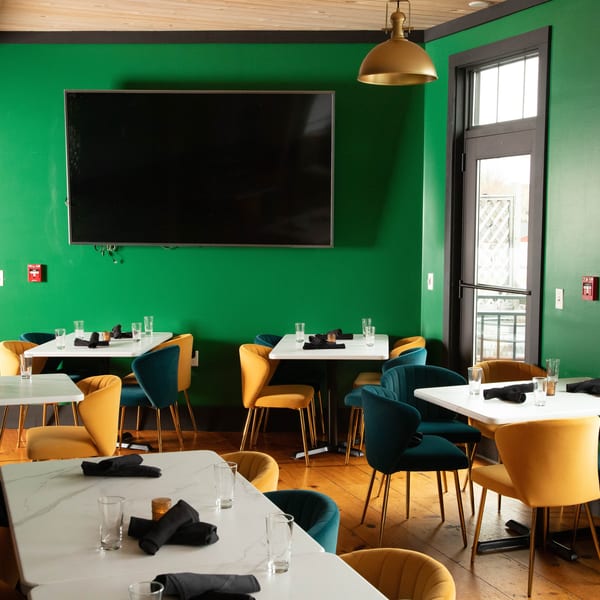 Restaurant dining room with green walls, wooden floors, white tables, and mustard yellow and teal chairs.