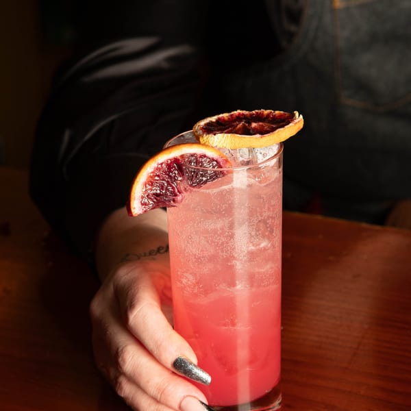 A hand with glittery nails holds a tall glass of pink cocktail garnished with blood orange slices.