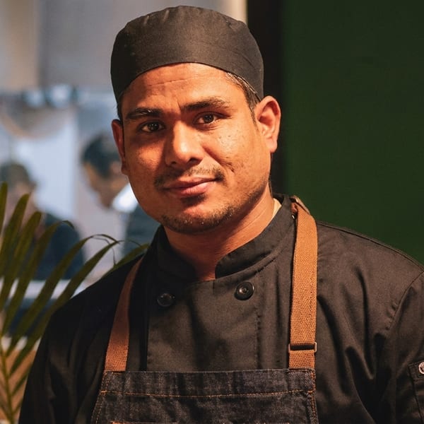 A chef in a black uniform and denim apron smiles at the camera.