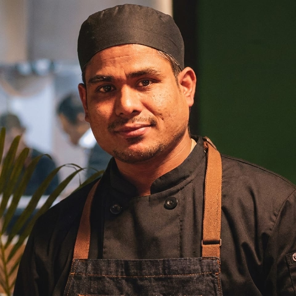 A chef in a black uniform and denim apron smiles at the camera.
