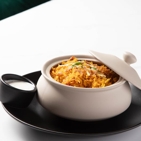 A clay pot of vibrant orange biryani with fried onions and green chilies, served with raita.