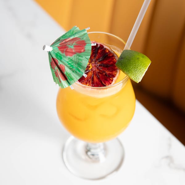 A vibrant orange cocktail in a stemmed glass, garnished with a lime wedge, blood orange slice, and a small umbrella.