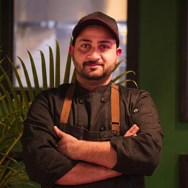 A chef in a black uniform and cap, with crossed arms, stands in a restaurant.