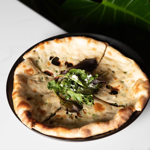 Round flatbread, cut into wedges, topped with green herbs, cheese, and a small salad.