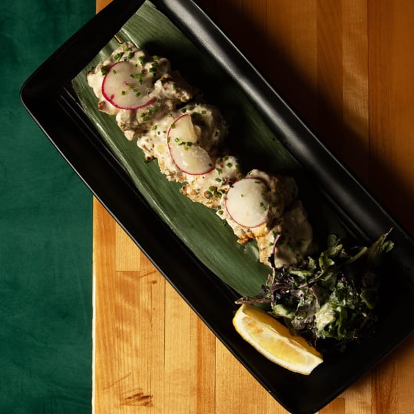Skewered chicken with creamy sauce, radish, chives, and a lemon wedge on a black plate.