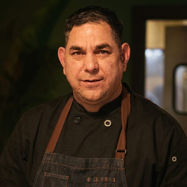 A male chef in a black uniform and denim apron stands in a restaurant kitchen.
