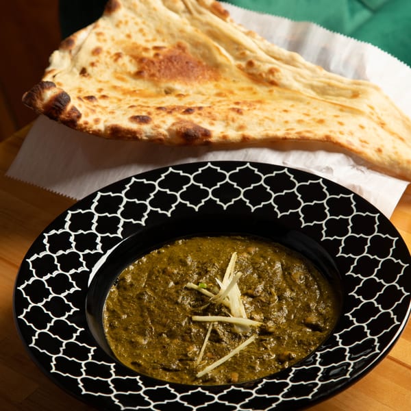 A black bowl of green curry with ginger slices, served with naan bread on a wooden table.