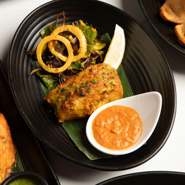 Tandoori fish with onion rings, lemon, and orange sauce on a black plate.