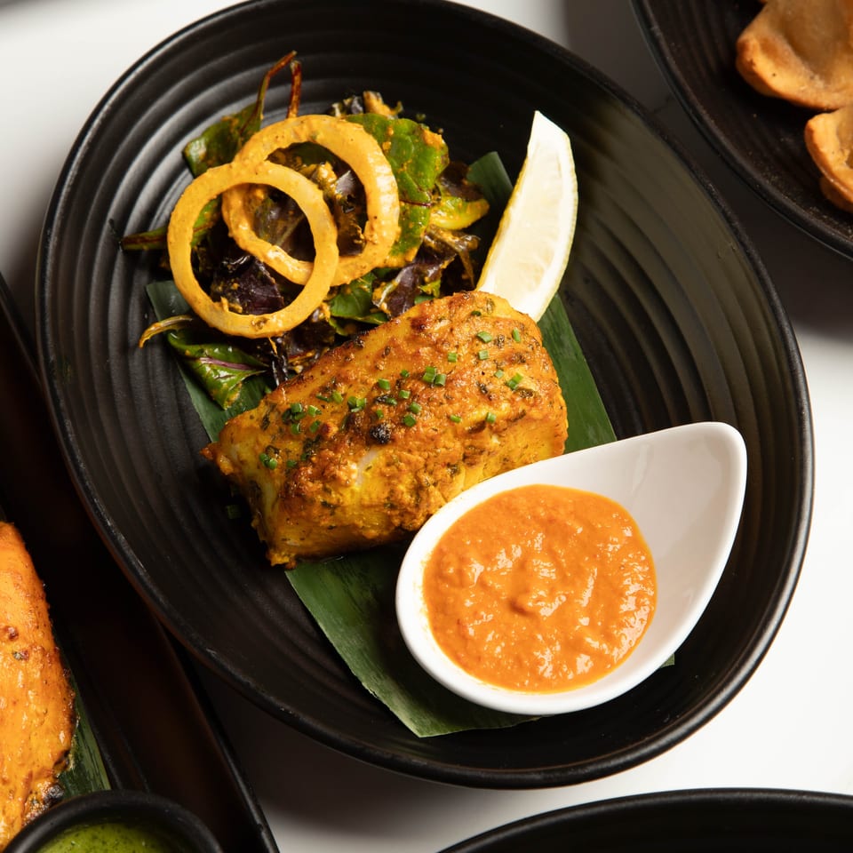 Tandoori fish with onion rings, lemon, and orange sauce on a black plate.