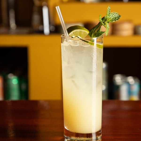 Tall glass of light yellow cocktail with ice, lime, and mint on a wooden bar.