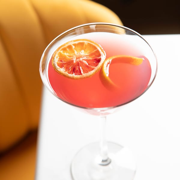 A vibrant pink cocktail in a martini glass, garnished with a blood orange slice and orange peel.