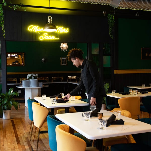 A server setting tables in a restaurant with green walls, neon sign, and colorful chairs.