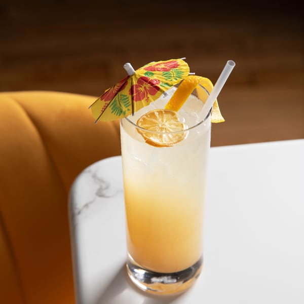 Tall glass of yellow cocktail with ice, lemon, orange peel, straw, and paper umbrella.