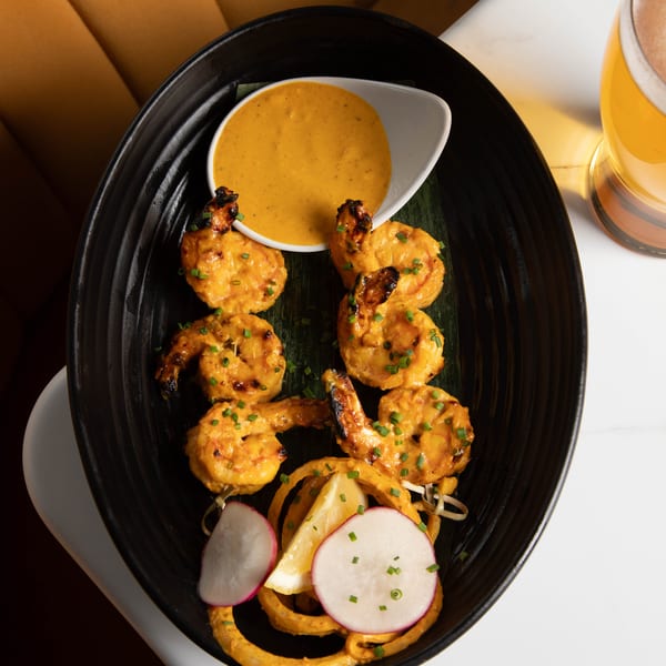 Grilled shrimp skewers with sauce, radishes, and lemon on a black plate with a beer.
